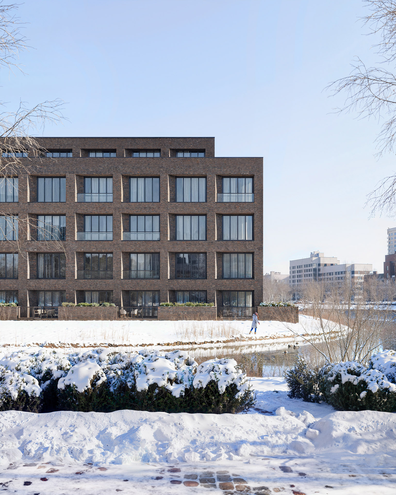 Rendering of a modern brick residential building in a snowy landscape with a person walking by a frozen pond