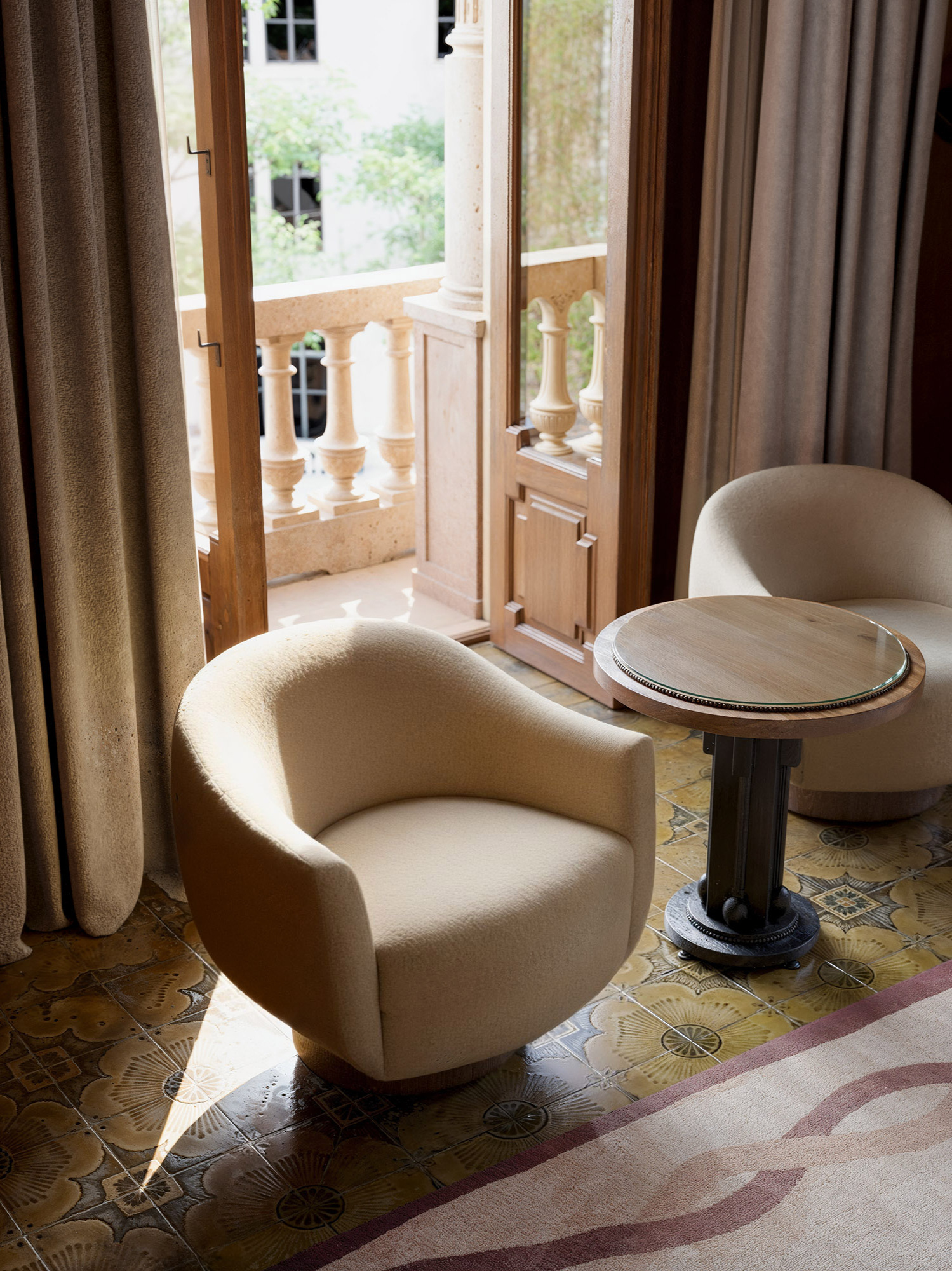 Two modern beige lounge chairs and a round table by an open window, with natural light highlighting patterned floor tiles and a warm-toned built-in bar in a sophisticated interior