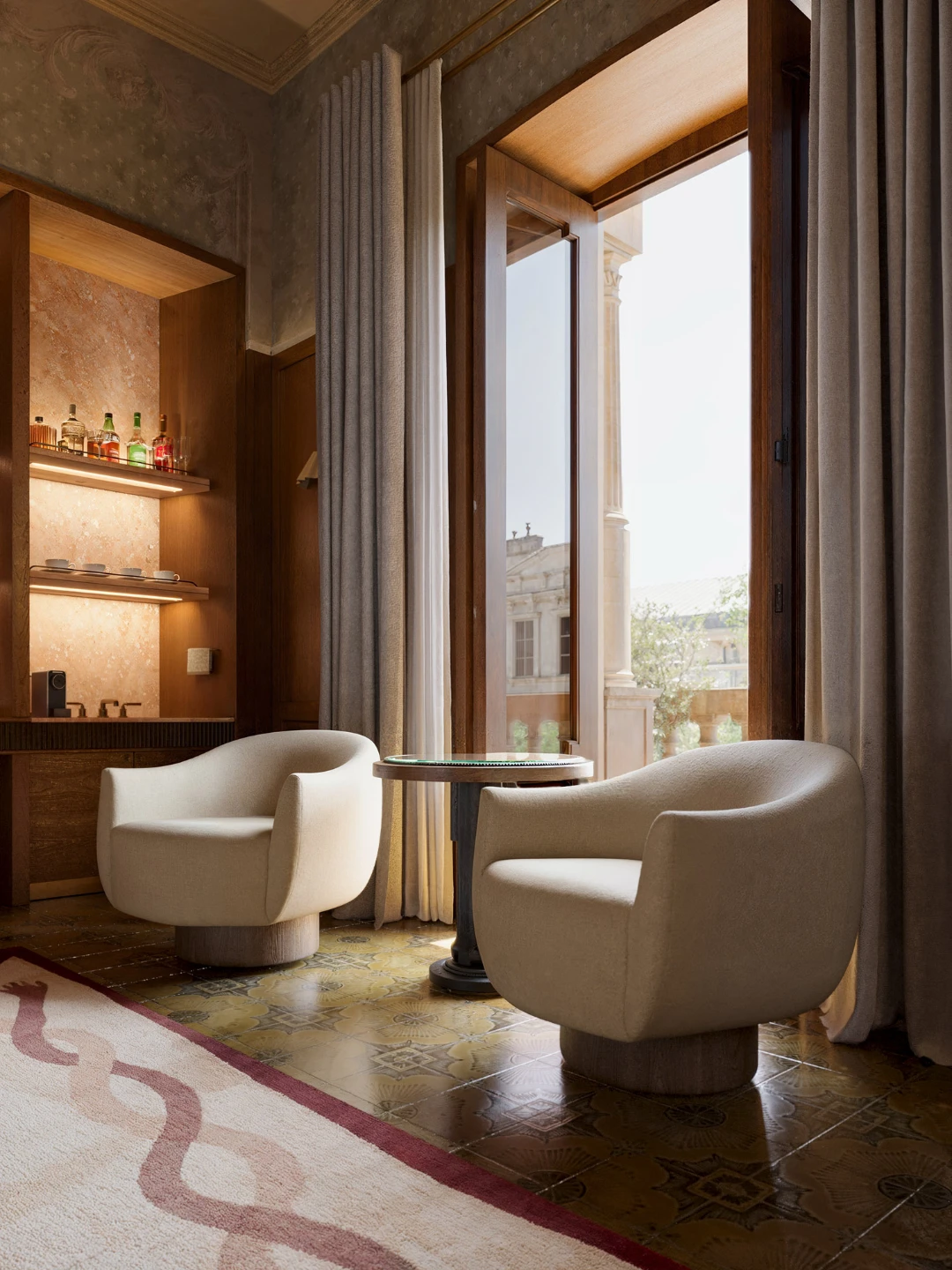 Luxury hotel suite sitting area with cream bouclé tub chairs, backlit marble bar niche, and open French window overlooking classical European facade — architectural interior rendering