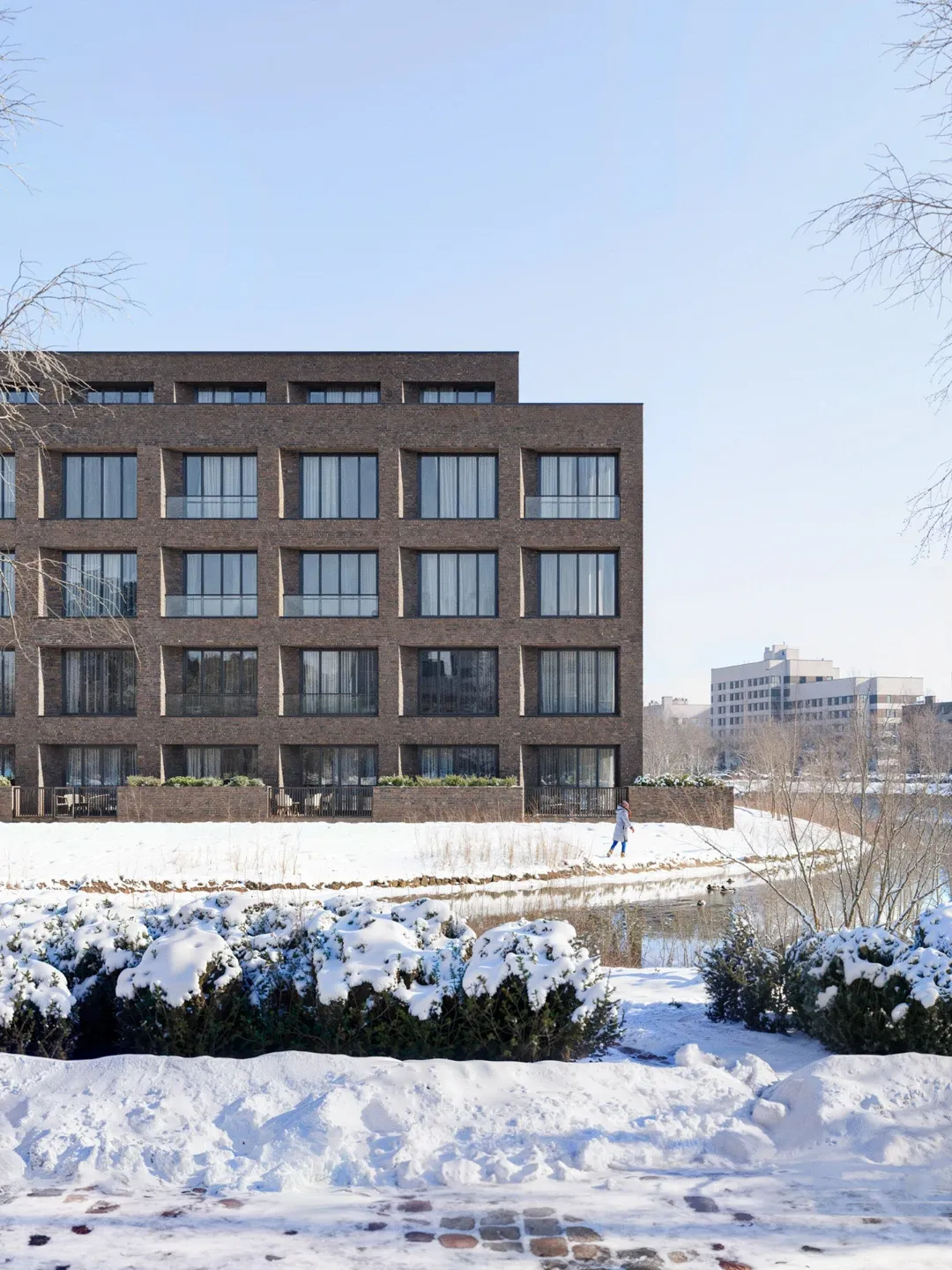 Dark engineering brick residential apartment building with arched window reveals overlooking snow-covered riverside park in winter — architectural exterior rendering